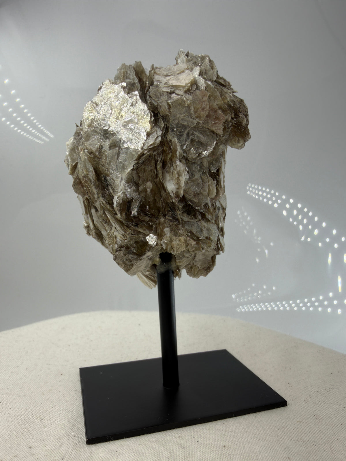 Silbrig glänzender Muskovit-Glimmer auf einem schwarzen Metallständer zur dekorativen Präsentation.