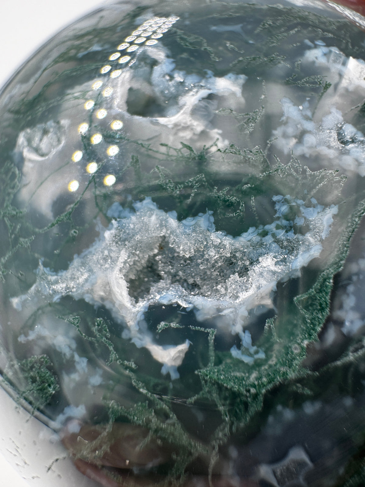 Eine hochglanzpolierte Moosachat-Kugel mit tiefgrünen, pflanzenähnlichen Strukturen in einer teils transparenten Matrix.