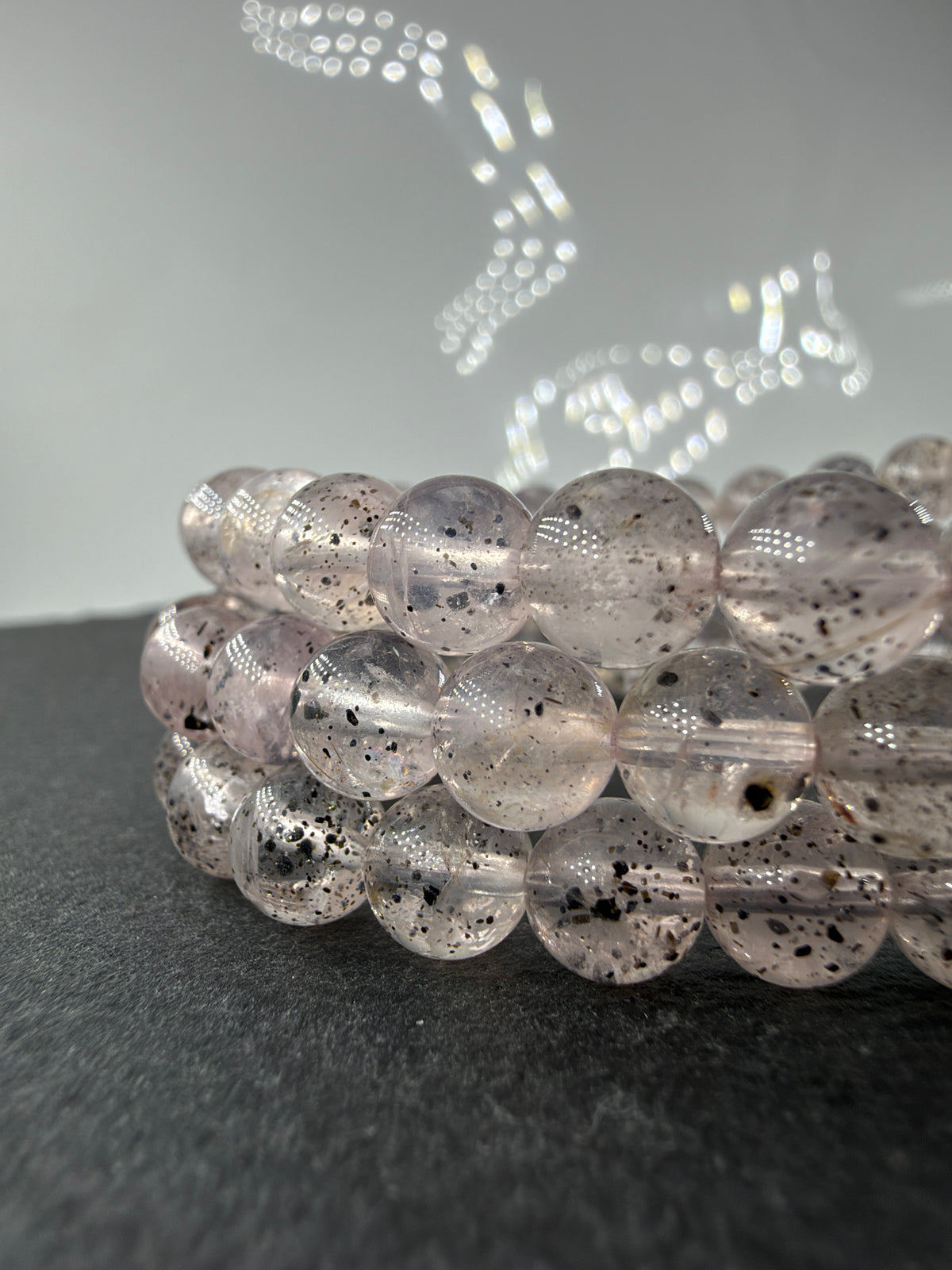 Rosenquarz-Hämatit-Armbänder RM22 mit zartrosa, durchscheinenden Kugelperlen und dunklen Einschlüssen auf dunklem Untergrund.